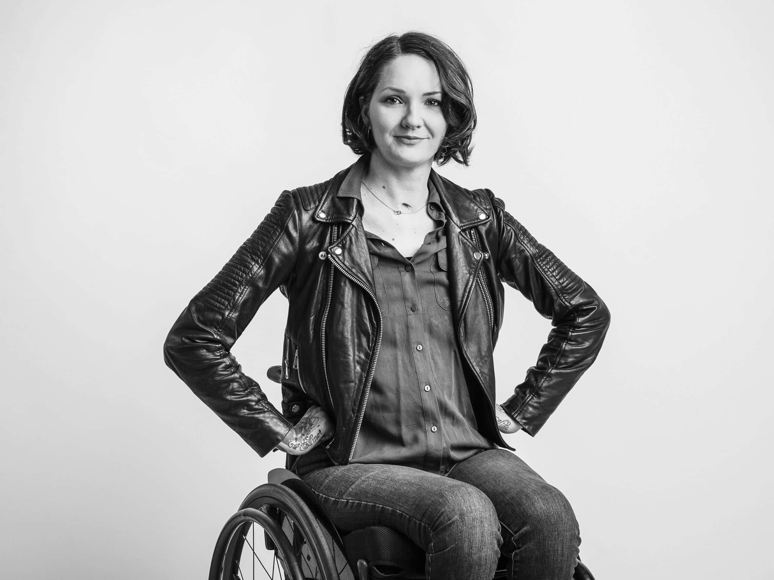 Svartvit porträtt av Yasmin Nilson som tittar mot kameran. Hon sitter i rullstol och håller händerna på höfterna. Hon har på sig skjorta, svart skinnjacka och jeans.
