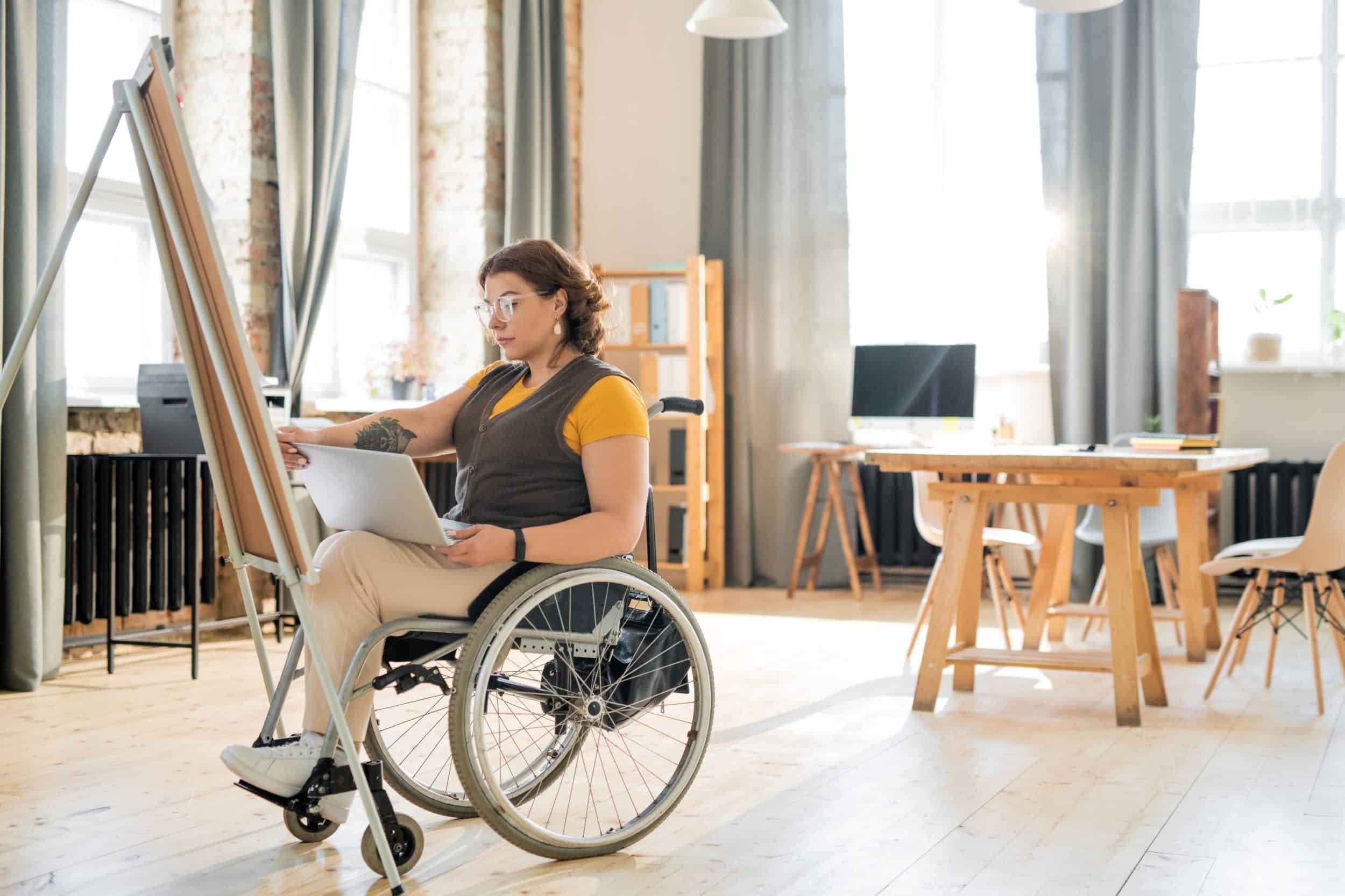 Person i rullstol sitter i ett rum och har en dator i knäet. I bakgrunden syns vacker inredning och långa gardiner.