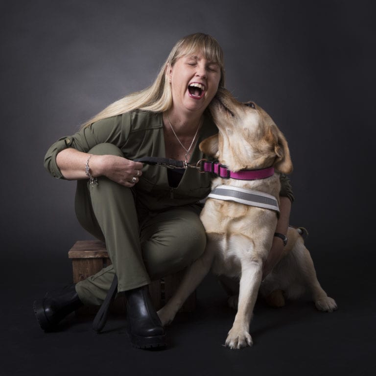 Anna Bergholtz sitter på huk och gapskrattar med sin ledarhund bredvid som slickar henne i örat. Anna har en grön byxdress på sig och långt blont hår. Ledarhunden är en krämfärgad labrador.
