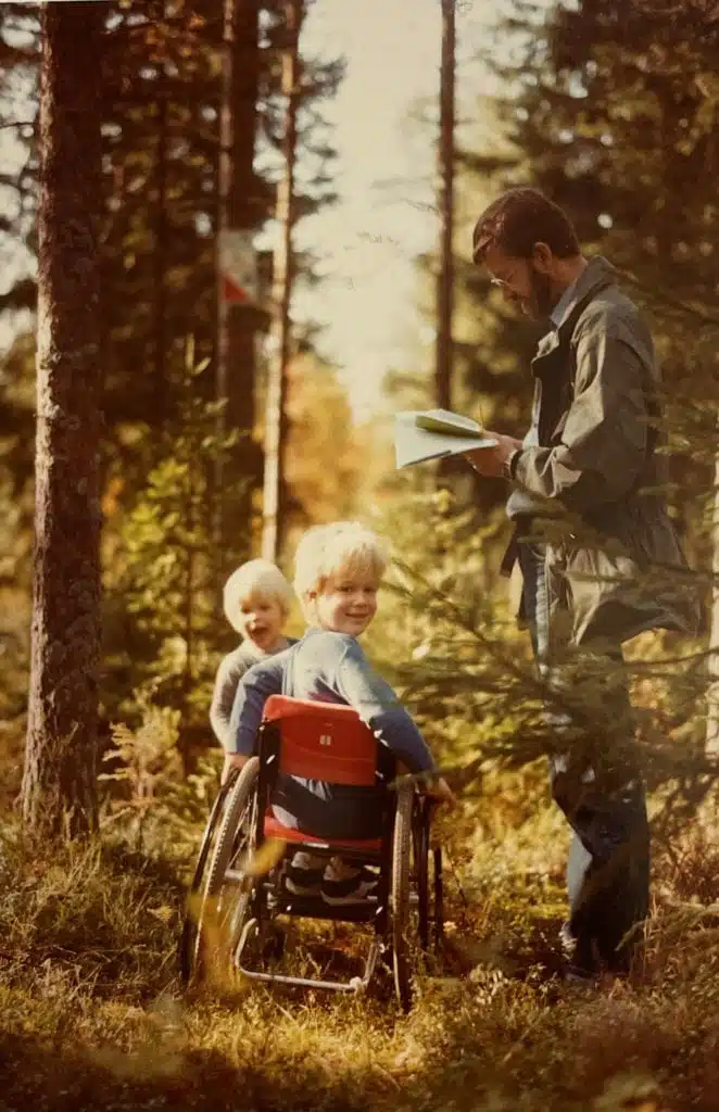 Staffan när han 5 år gammal använder en av sina första rullstolar för barn. Han rullar i skogen med sin far och lillebror. Staffan är vänd mot kameran och ler, lillebror kikar fram skrattande och pappan står och läser i en karta. 