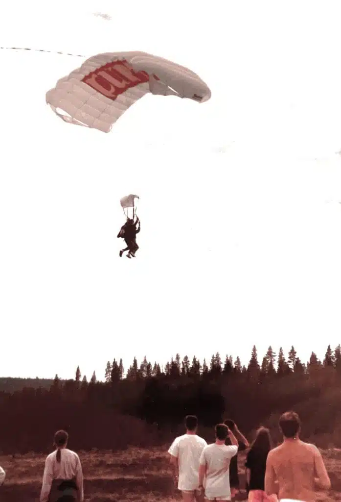 Staffan provar på tandemfallskärmshopp i Västerbotten som tonåring. Staffan seglar ned i fallskärmen och människor står nedanför och tittar.