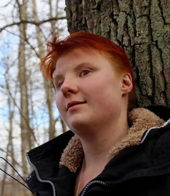 Desideria Jungelin. Hon har kort rött hår, svart jacka med beige pälskrage. I bakgrunden syns träd och blå himmel.