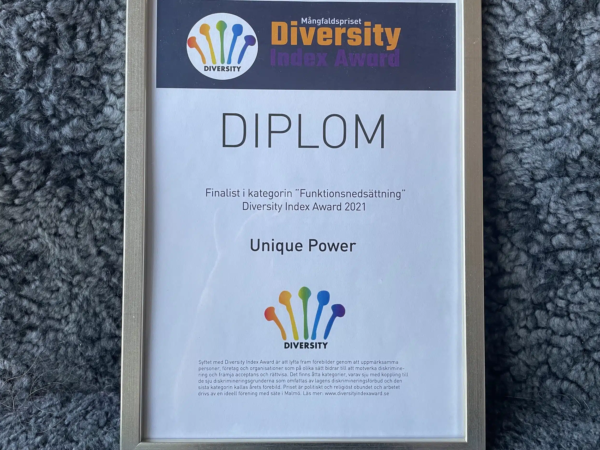 Diplom från Diversity Index Award. Ett diplom liggandes mot grå bakgrund. Längst upp logotyp med 5 färgglada spröt. "Finalist i kategorin "Funktionsnedsättning" Diversity Index Award 2021".