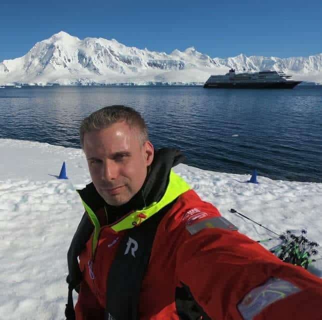 Rasmus Isaksson selfiefoto. Han har kort mörkblont hår, en röd och neongul vinterjacka. I bakgrunden syns snö på marken, vatten och längre bort snötäckta bergskroppar samt ett fartyg.