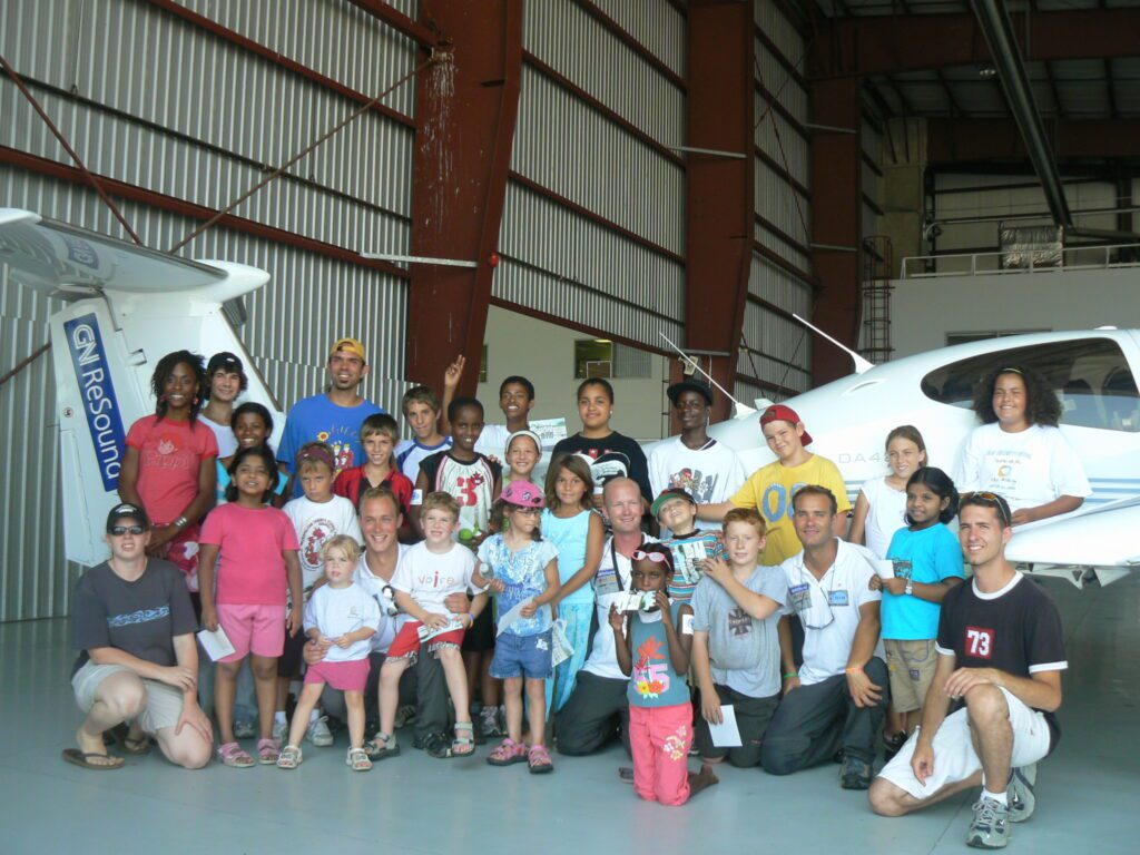En grupp på cirka 20 personer är uppställda för gruppfoto framför ett litet vitt propellerflygplan i en hangar. Persomerna på bild är av olika ålder och etnicitet.