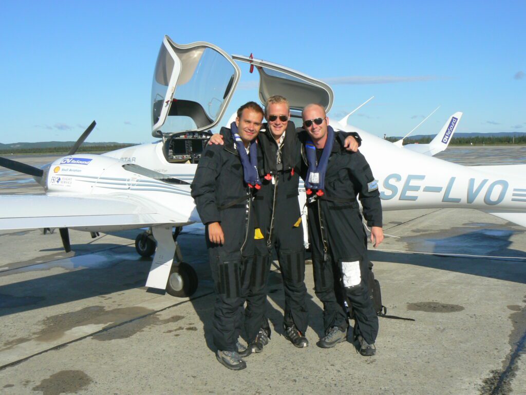 Johan står med två kollegor iklädda svarta overaller. Bakom syns ett par vita små propellerflygplan på ett öppet flygfält en sommardag.