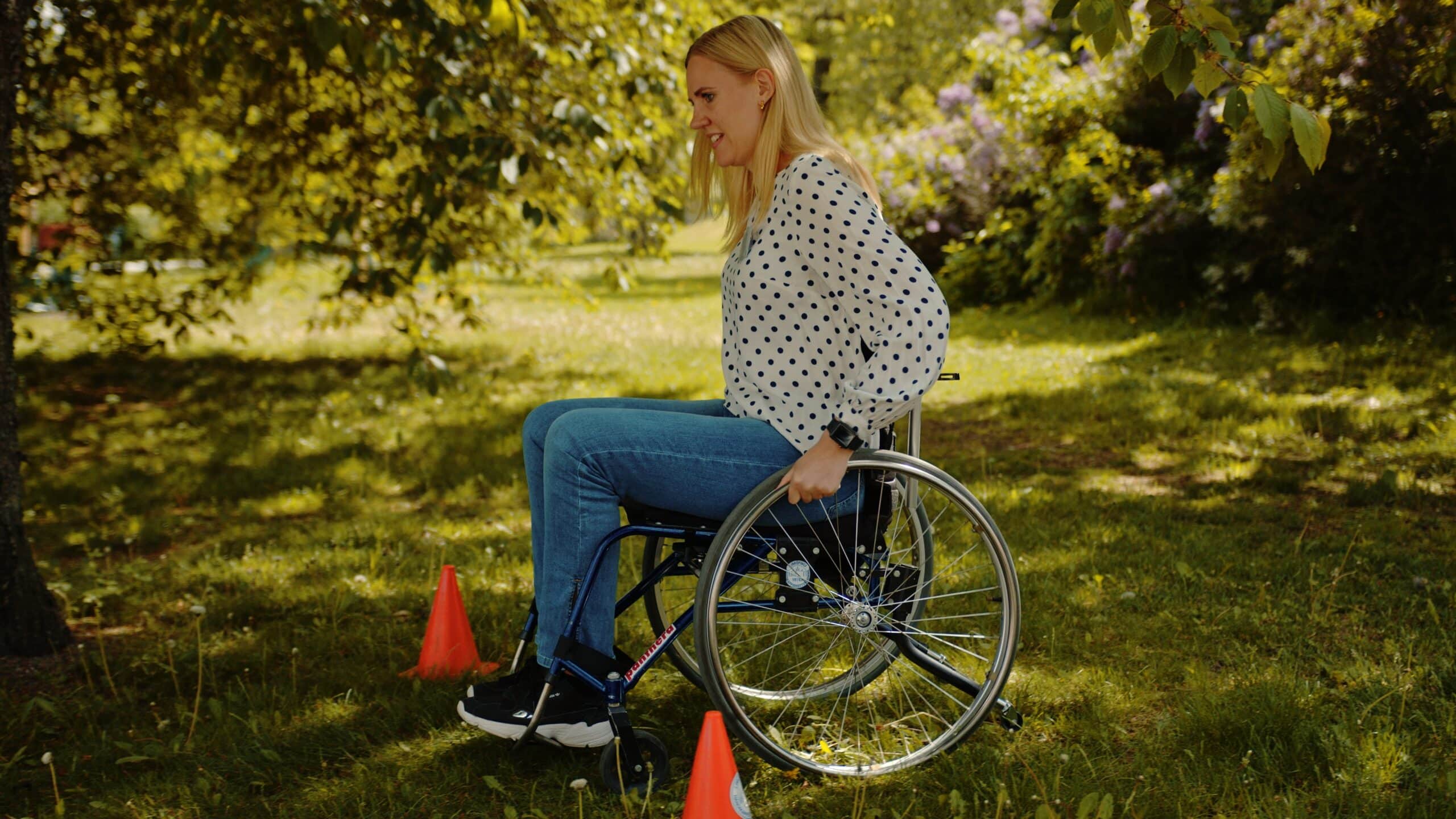 Blond kvinna sitter i rullstol i grönska och övar på en hinderbana med koner. Hon har vit tröja och blåa jeans, rullstolen är svart sett från sidan.