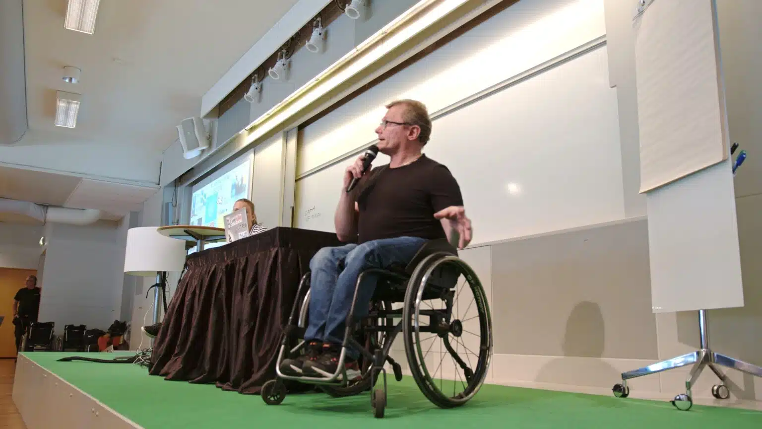 Föreläsare Lars-Göran Wadén och Anna Bergholtz på scen. Lars-Göran sitter i sin rullstol och bakom syns Anna vid en laptopskärm. På väggen en whiteboard.