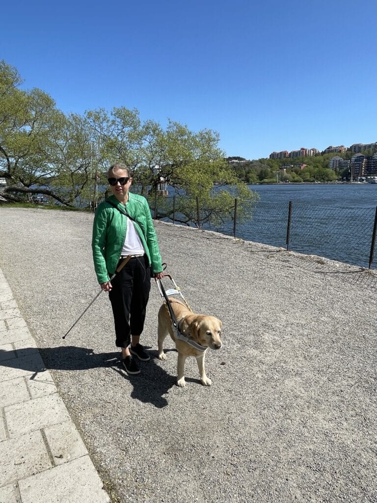 Anna Bergholtz och hennes ledarhund Djurgården 2023. Anna står utomhus på en grusväg med vatten och gröna träd i bakgrunden. Hon har på sig en grön jacka, vit tröja, svarta byxor och låga sommarskor. I ena handen håller hon en vit käpp. Med andra handen håller hon i sin ledarhund, en ljus labrador.