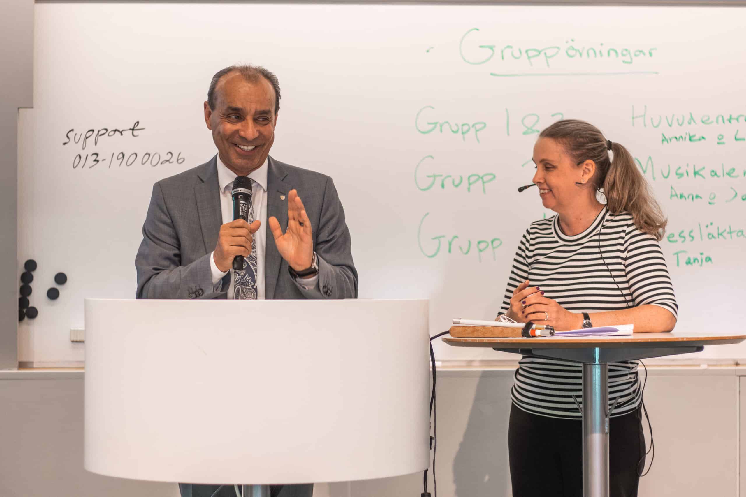 Man och kvinna pratar i föreläsningsrum framför en whiteboard. Mannen har grå kostym, kort gråsvart hår och håller i en mikrofon. Kvinnan har svartvitranding tröja och headset.