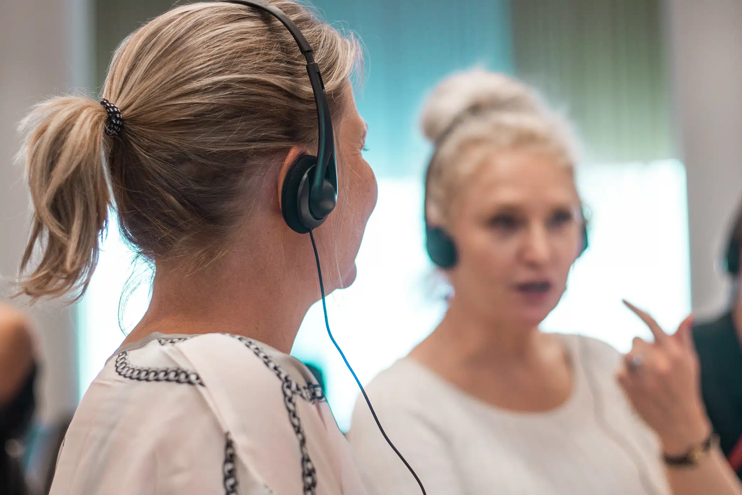 Två kvinnor sitter i sal med hörlurar under en hörselövning. Båda har vita tröjor och ljusblont hår och tittar mot varandra engagerat.