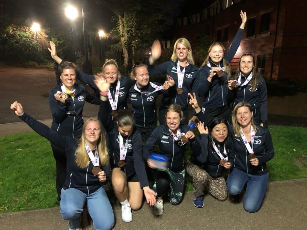 Sveriges lag med 11 glada tjejer i mörka kläder, alla medaljerna runt halsen. Gruppen står utomhus vid gatlyktor vid en grässlänt på kvällen.