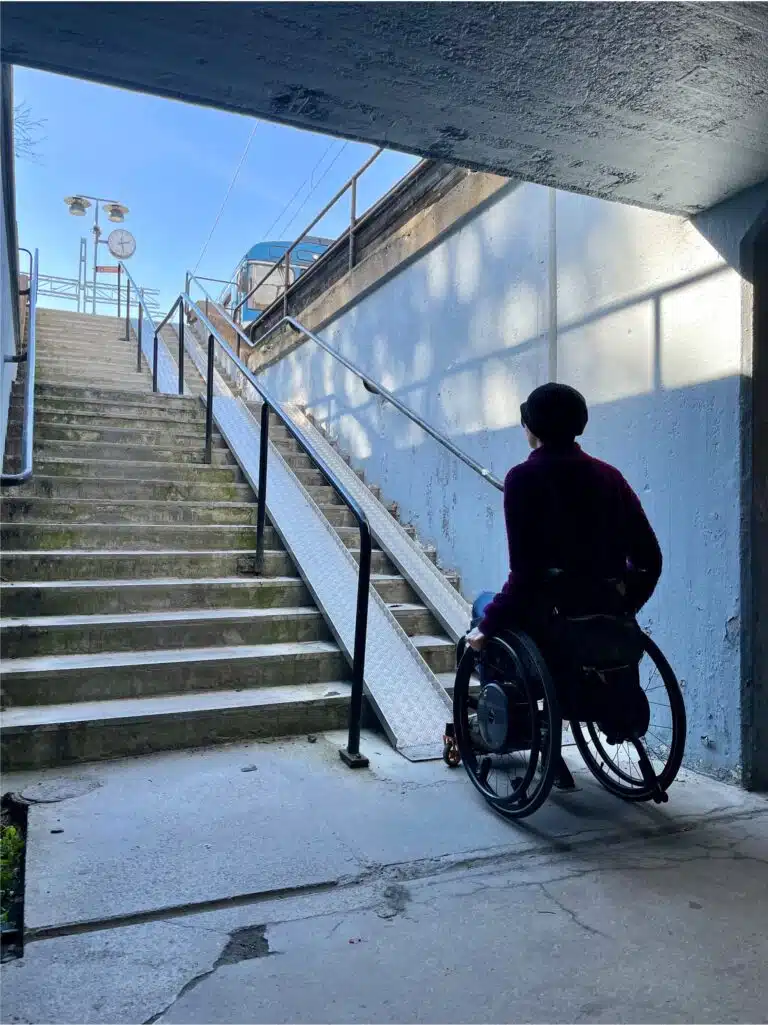 Siluett av kvinna i rullstol som sitter längst ned mot en trappa som har rampskenor på höger sida. Bakom syns taket i en mörk tunnel och ovanför trappan skymtar en spårvagnsperrong.