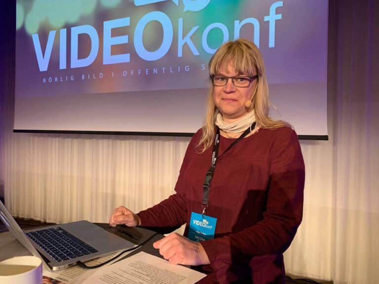 Helena på scen framför en overheadbild. Hon har långt ljust hår, glasögon och bär ett headset. Hon har på sig en mörk tröja, ljus scarf och står vid ett podium med en bärbar dator.