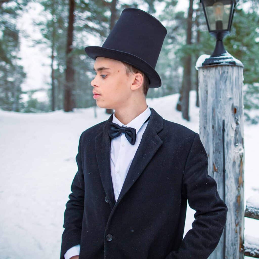 Viktor står intill ett trästaket på snö tittar åt sidan. Han har kort brunt hår, klädd i en svart hatt, elegant jacka och fluga ovanpå en vit skjorta. I bakgrunden syns skog och vit snö.