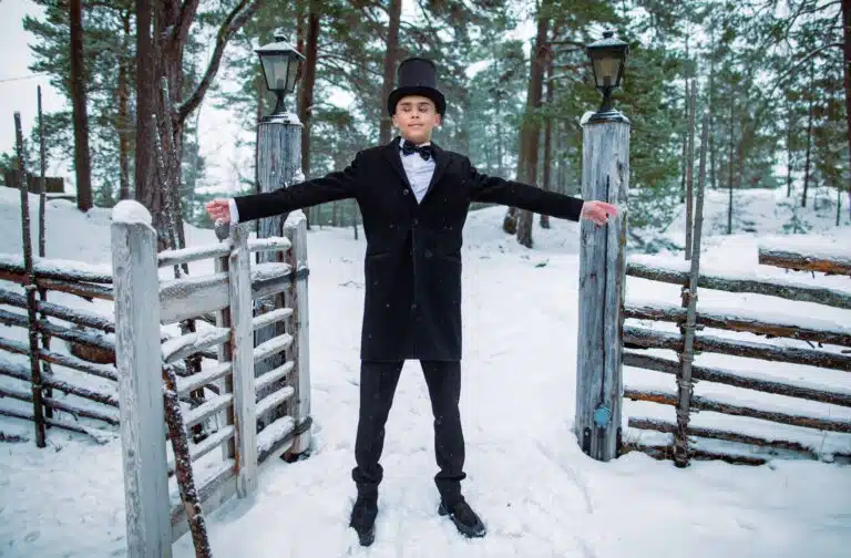 Viktor står med utsträckta armar på en väg med snö. Han har kort brunt hår, klädd i en svart elegant jacka, svart hatt, finskor och fluga ovanpå en vit skjorta. I bakgrunden syns ett trästaket och skog.