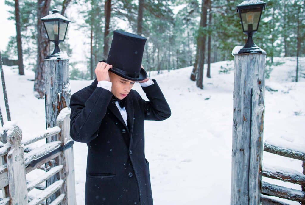 Viktor står på snö och håller om en svart hatt på huvudet. Han har kort brunt hår, klädd i en svart elegant jacka och fluga ovanpå en vit skjorta. I bakgrunden syns ett trästaket, snö och skog.