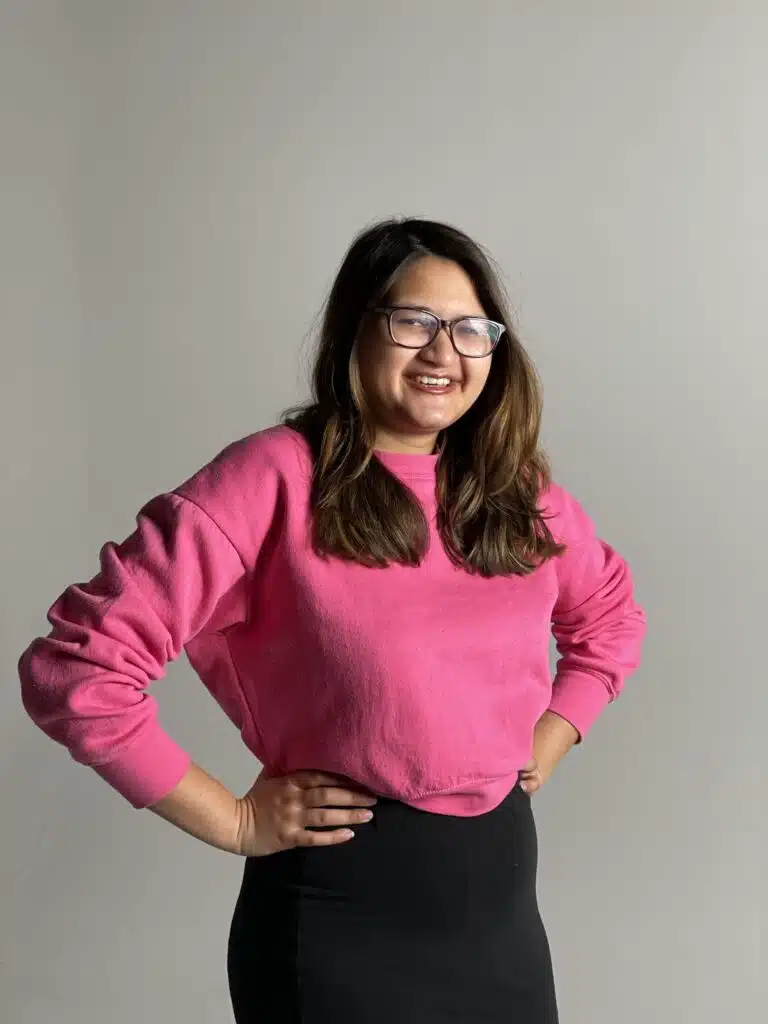 Tania Gazi i fotostudio med grå bakgrund. Hon har svart långt hår, glasögon, ler mot kameran och står med händerna mot midjan iklädd en rosa tröja och svart kjol.