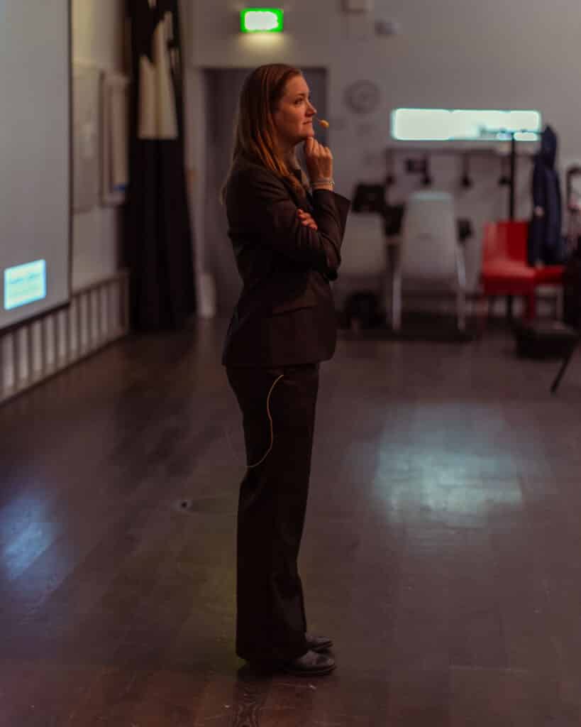 Susanna Cederquist står framför en projektorduk med headset. Hon har långt blont hår, klädd i en svart kavaj, svarta byxor, svarta skor och tittar ut mot sidan av fotot. 