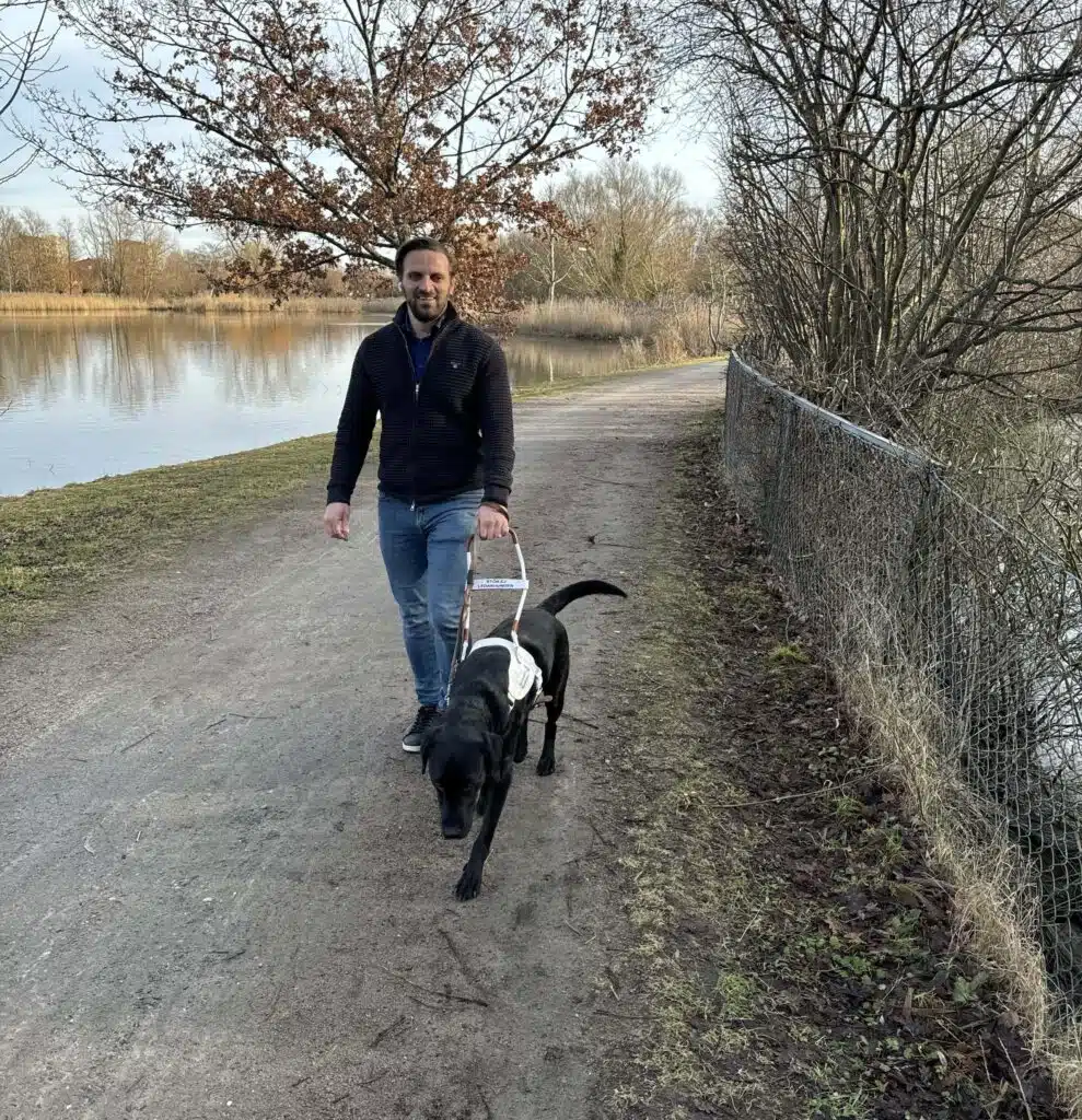 Fatmir går med sin ledarhund Komet går på en grusväg intill en sjö med buskage runtom. Fatmir har kort brunt hår, skägg och ler. Han har på sig svart fleecejacka, blåa jeans och mörka sneakers. I sele håller han sin mörka ledarhund som går just framför.