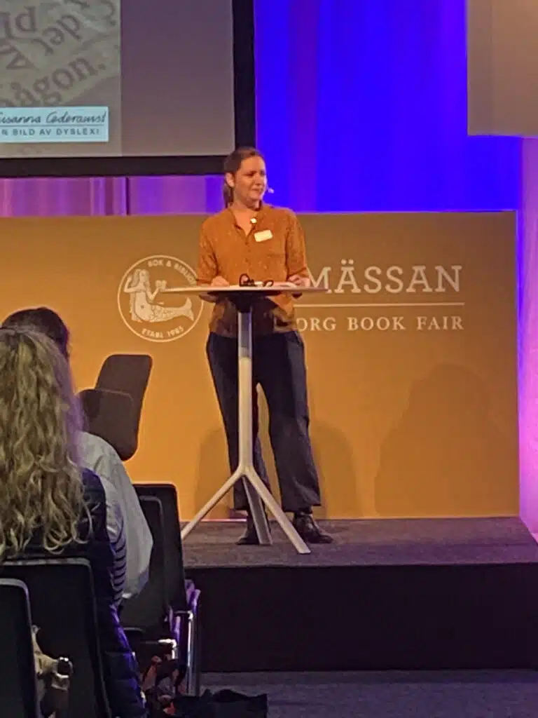 Susanna Cederquist föreläser vid podium på bokmässan. Hon har mörkt brunt långt hår, headset och klädd i en ljusbrun skjorta, mörka byxor och skor. Hon ler och tittar ut mot åhörarna.