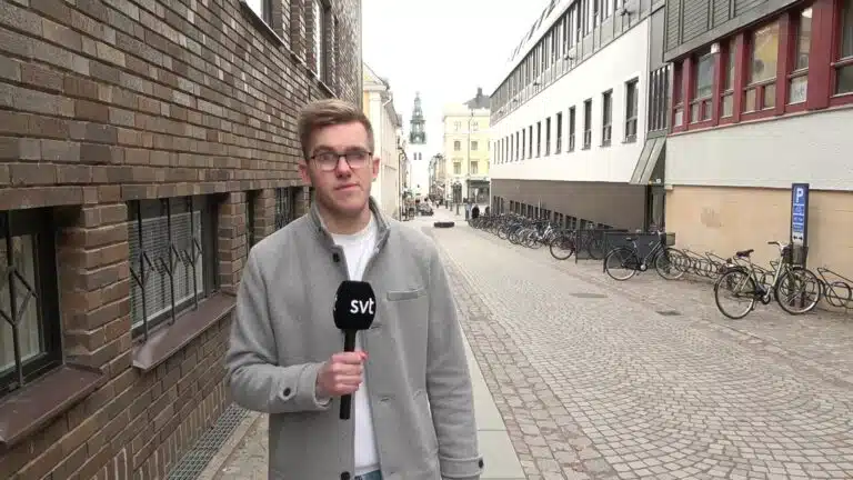 Martin Klevelid står utomhus i en gränd och håller i en mikrofon med texten SVT. Han har kort ljust hår, glasögon, en vit t-shirt under en grå jacka