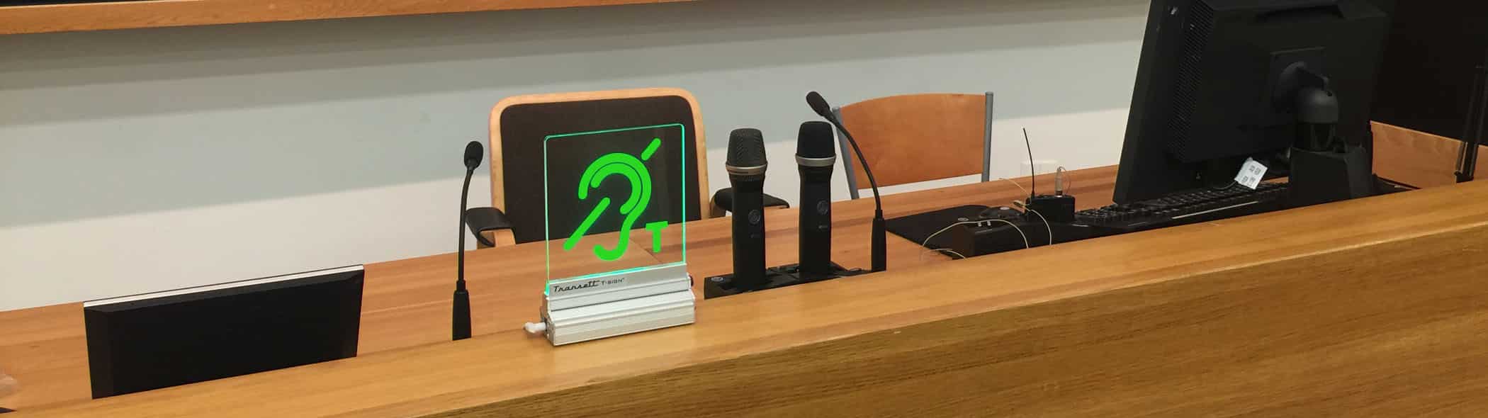Närbild av T-sign monterat på ett bord intill några mikrofoner, datorskärmar och stolar i en föreläsningsmiljö