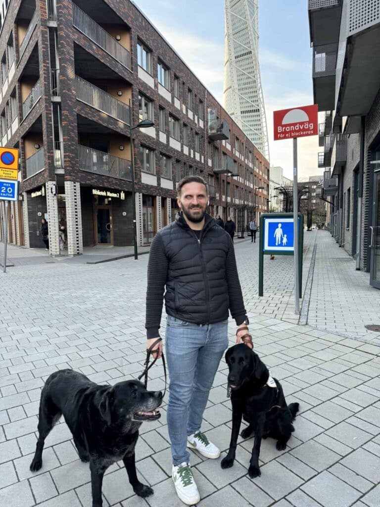 Fatmir står ute på ett torg nedanför Turning Torso i Malmö. Han håller i två svarta ledarhundar i varsitt koppel. Fatmir har kort brunt hår och skägg samt är klädd i svart jacka, blå jeans och vita sneakers.