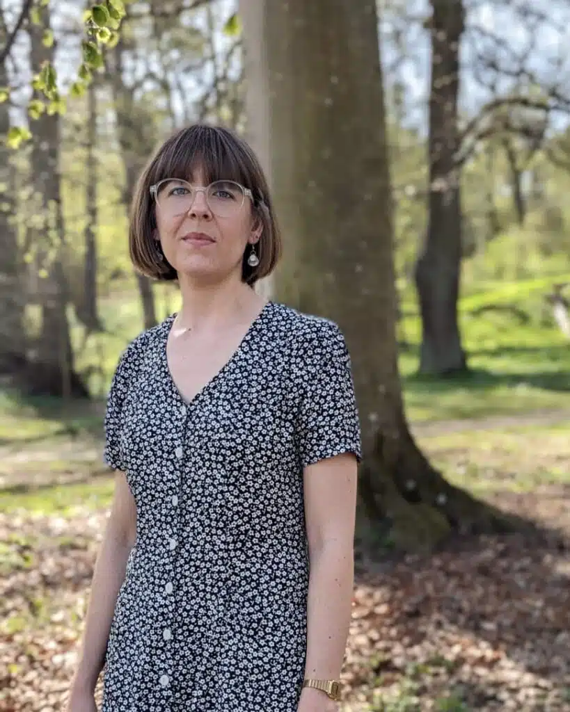 Ida Hansson står i skogen iklädd en vitprickig/svart klänning. Hon har halvlångt brunt hår, glasögon, tjusiga örhängen och en guldklocka på sig.