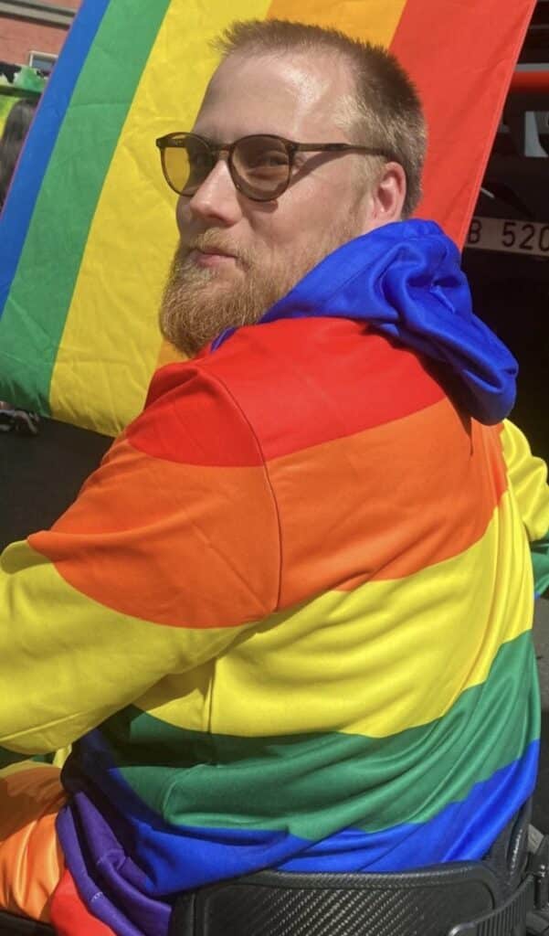 Oscar Sjökvist har ljust kort hår och skägg samt glasögon. Iklädd regnbågsfärgad tröja sitter han i sin rullstol utomhus under en flagga med samma färger.