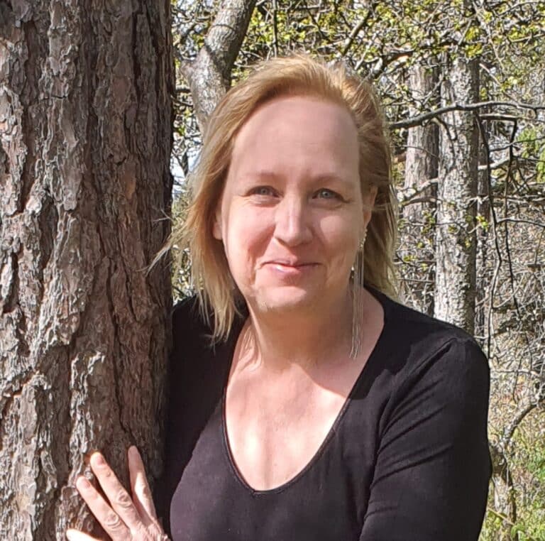Jenny Ström i skogsmiljö. Hon har långt blont hår ler och har långa örhängen på sig. Hon är klädd i en svart tröja och lutar sig mot ett träd.