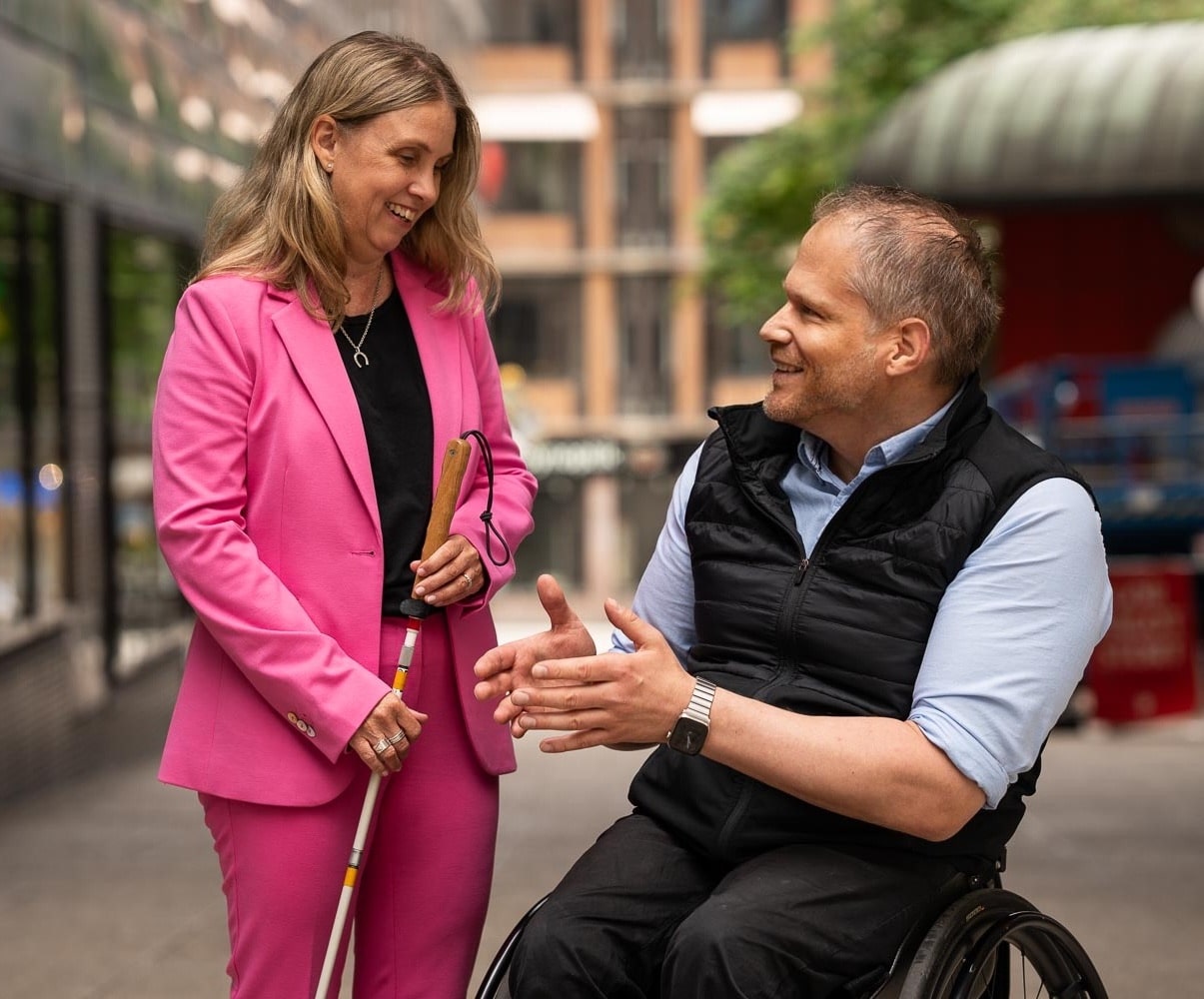 Anna och Staffan står utomhus och pratar med varandra. Staffan sitter i rullstol klädd i en svart väst och skjorta. Anna har en rosa kavaj och och byxor och håller i en vit käpp.