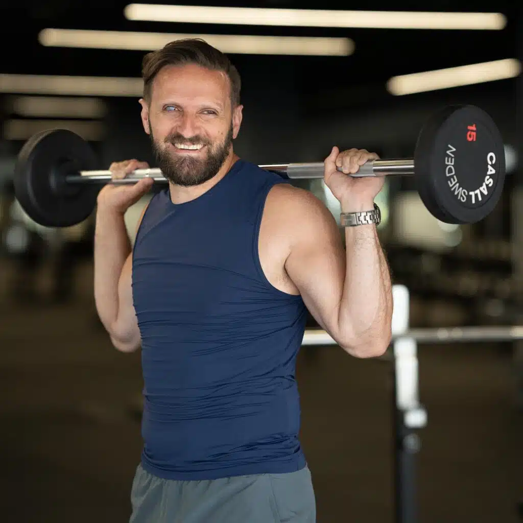 Fatmir Seremeti stående med en skivstång på axlarna klädd i blått linne, shorts och metallklocka. Han har brunt kort hår och brunt skägg med mustasch.