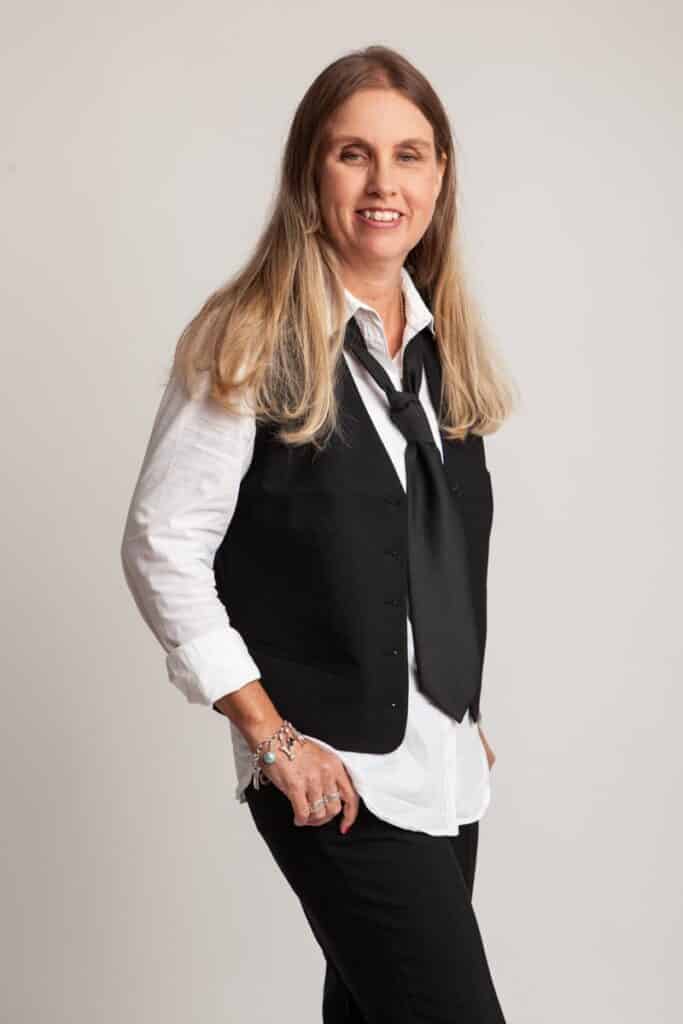 Profilbild på Anna. Hon står i fotostudio med långt blont hår, klädd i svart väst, vit skjorta, slips, svarta byxor och tittar mot bilden med ett leende.