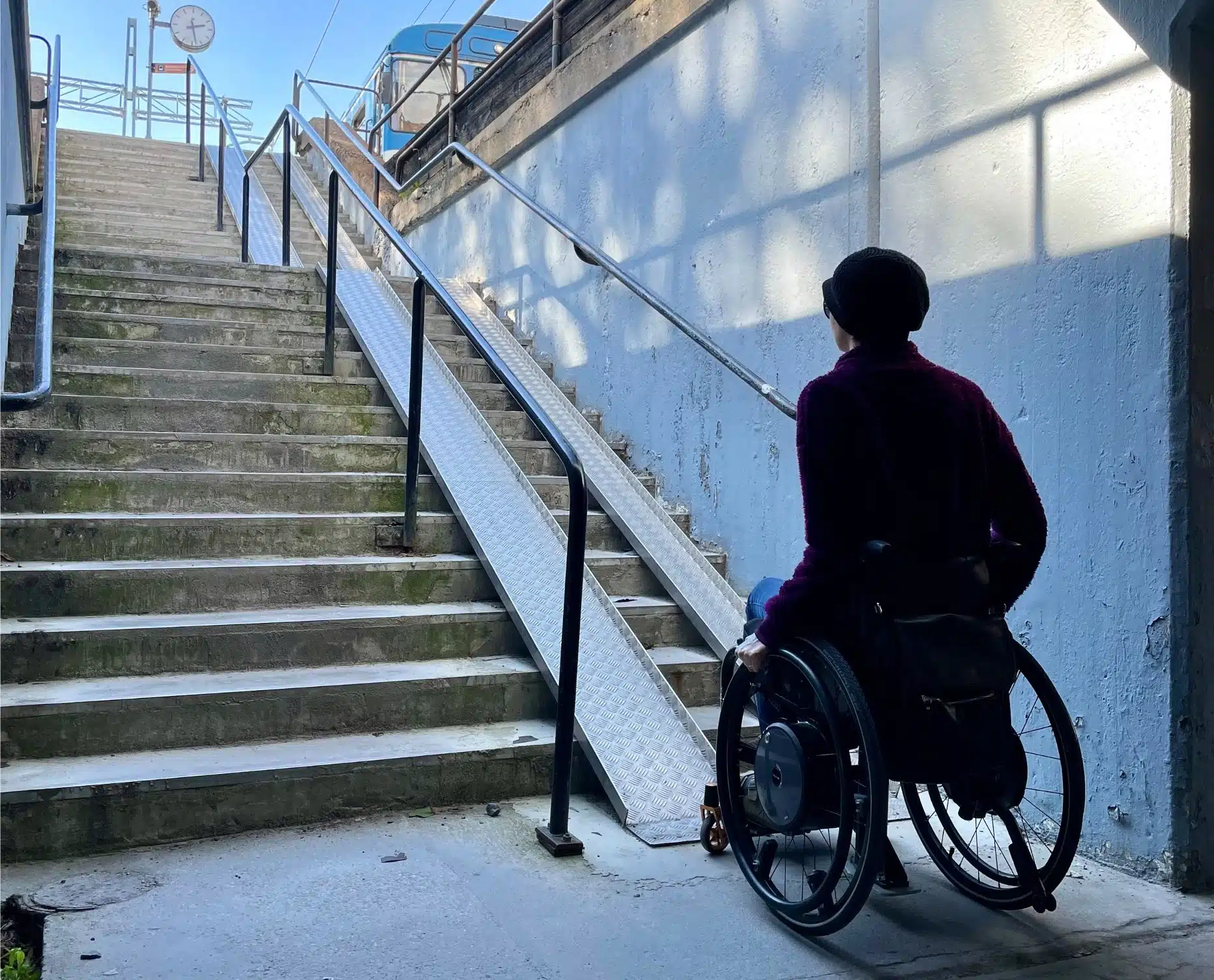 Siluett av kvinna i rullstol som sitter längst ned mot en trappa som har rampskenor på höger sida. Bakom syns taket i en mörk tunnel och ovanför trappan skymtar en spårvagnsperrong.