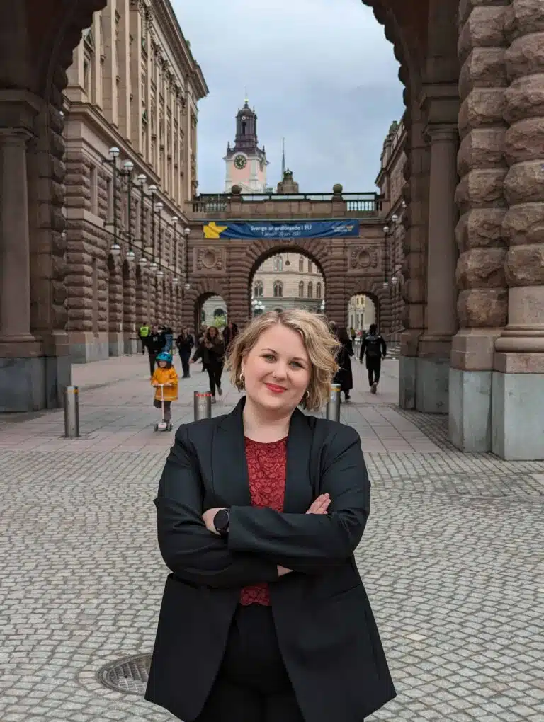 Johanna Forsberg står med armarna i kors på ett torg framför en portöppning mellan två byggnader. Hon har kort blont hår, rött läppstift och klädd i svart kavaj.