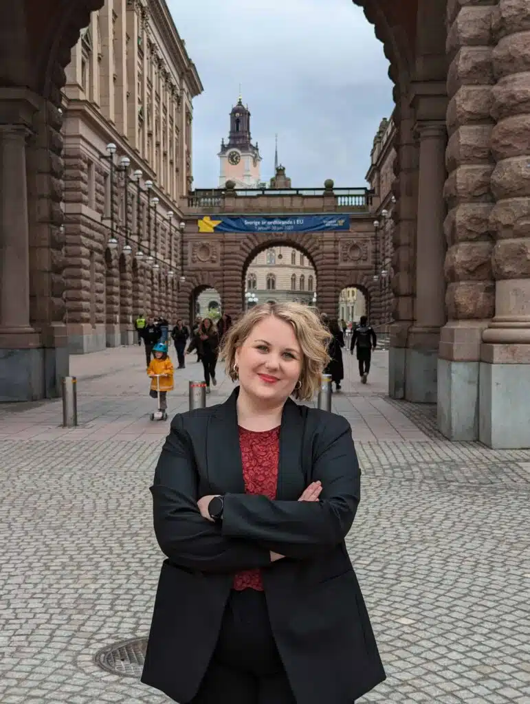 Johanna Forsberg står med armarna i kors på ett torg framför en portöppning mellan två byggnader. Hon har kort blont hår, rött läppstift och klädd i svart kavaj.