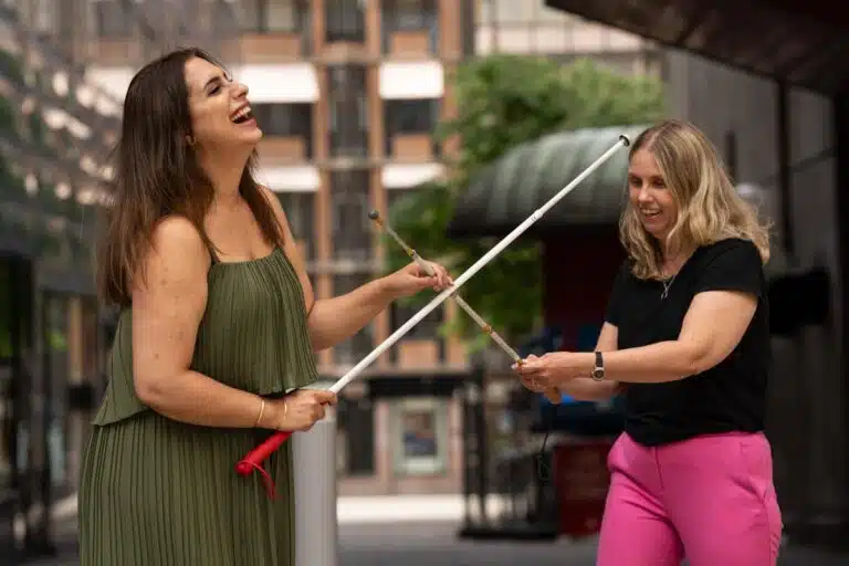 Parimah och Anna fäktas med varsin vit käpp utomhus på ett torg, Parimah har långt mörkbrunt hår och är klädd i en grön klänning. Anna har långt blont hår, svart t-shirt och rosa byxor.