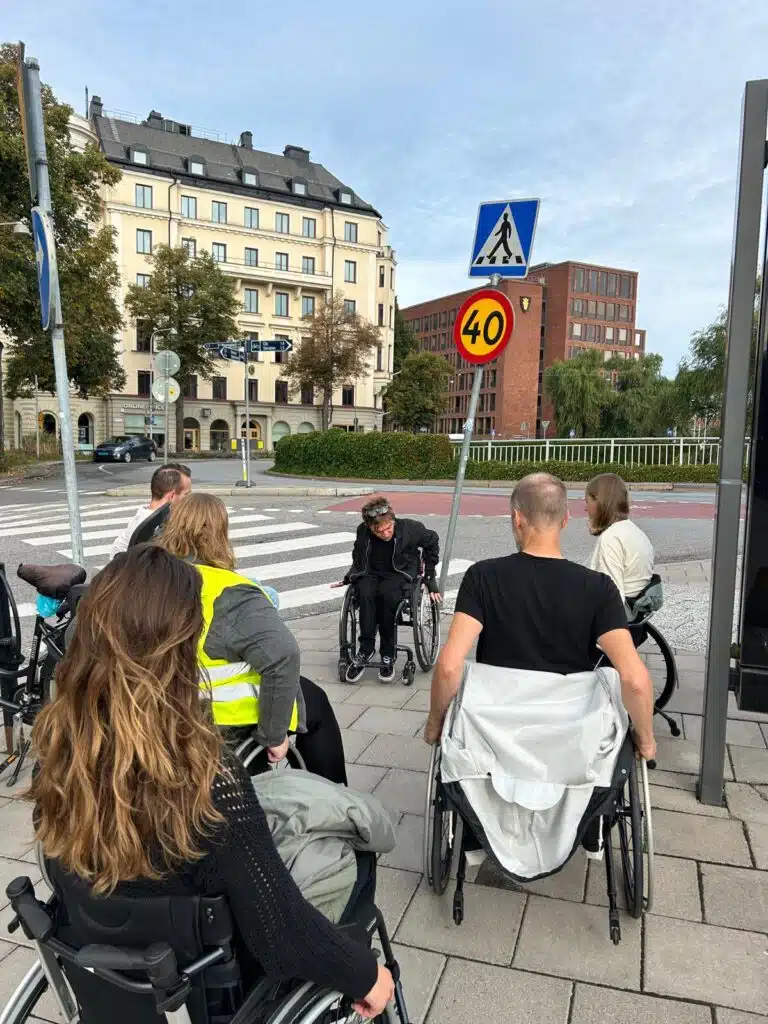 Fem deltagare är ute och rullar i fem rullstolar vid ett övergångsställe. Eddie instruerar hur man rullar