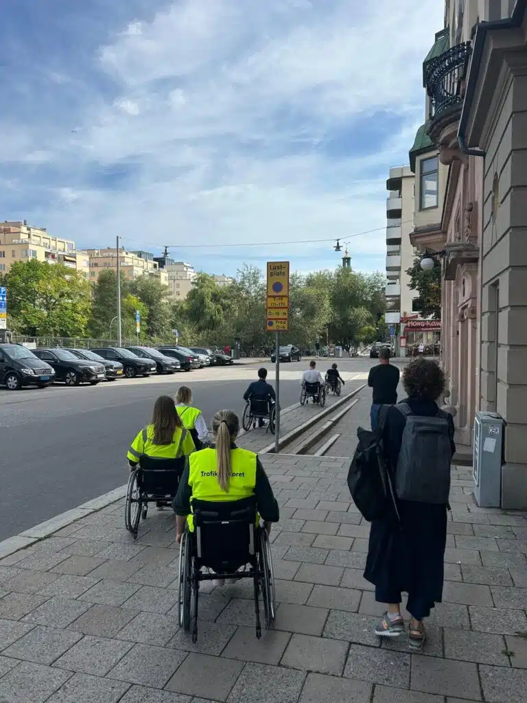 Fem deltagare i rullstol rullar på smal trottoar i nedförsbacke.