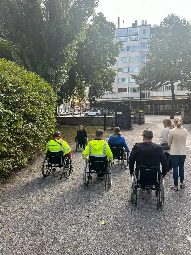 Fem deltagare i rullstol testar att rulla på grus. Eddie instruerar.