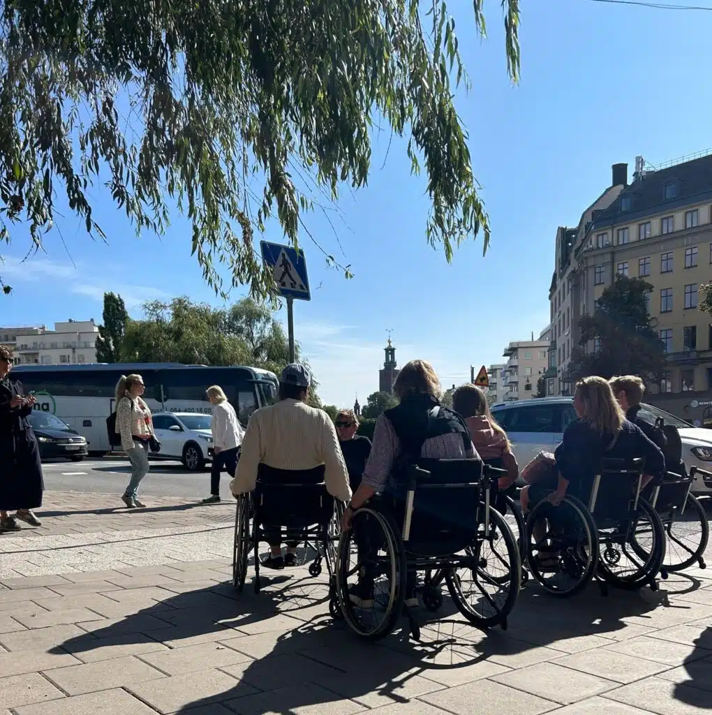 Fem deltagare i rullstol väntar på att rulla över ett övergångsställe i trafikerad väg.