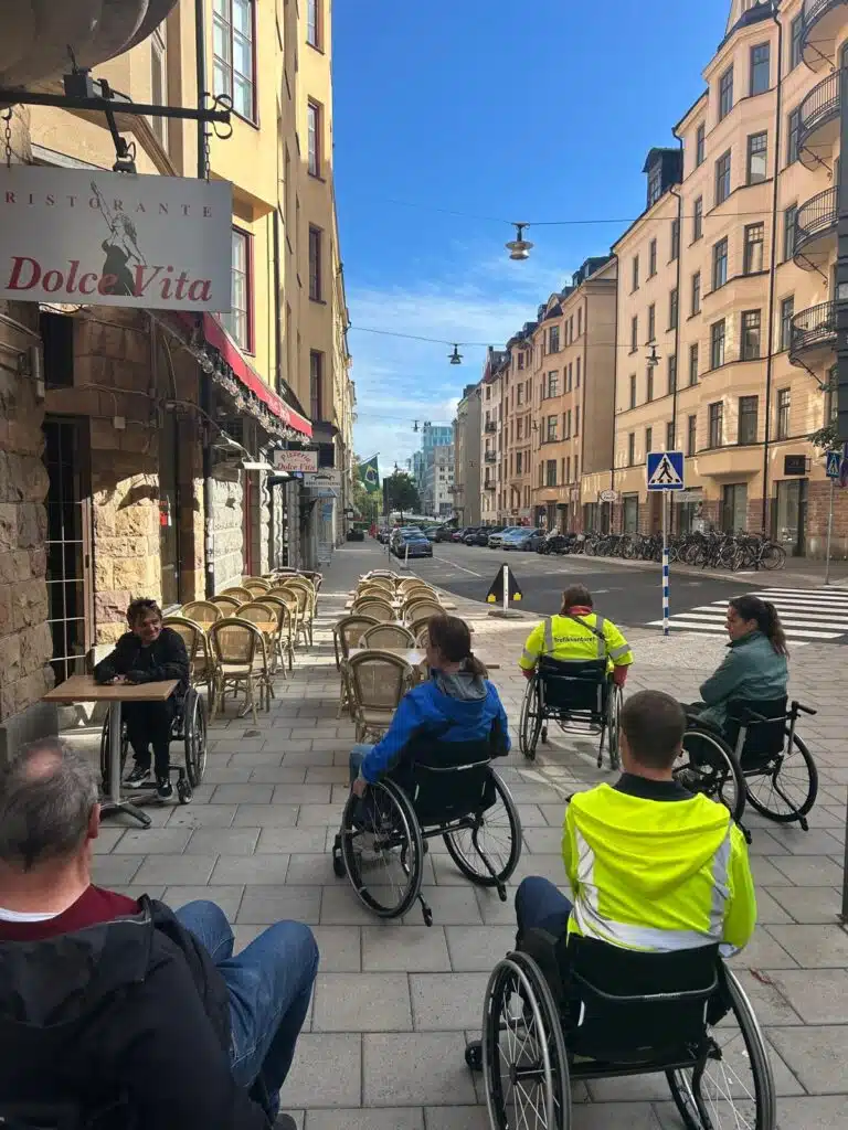 Fem personer, några i gula västar sitter i sina provrullstolar framför Eddie, en av våra konsulter som själv använder rullstol. Han visar hur man rullar intill ett cafébord med rullstol.