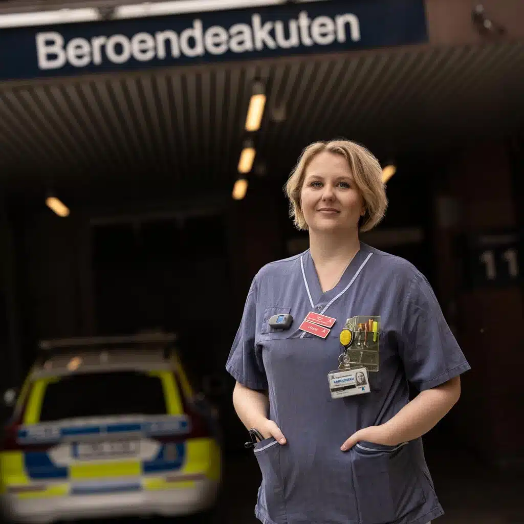 Johanna står med händerna i fickorna vid garageinfarten till en beroendeakutmottagning. Hon har axellångt blont hår och är klädd i sin blå arbetsklädsel med röd namnskylt på bröstet.