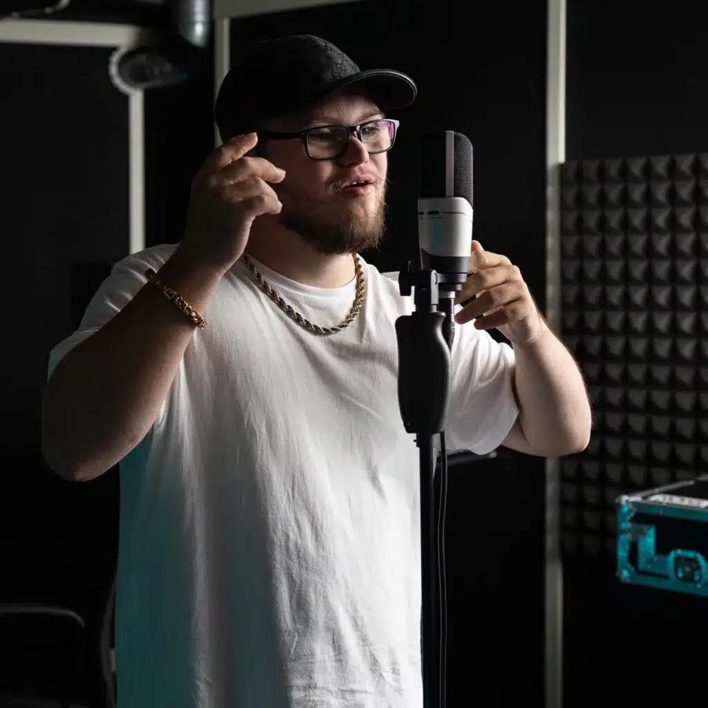 Leo står i en mörk studio intill en mikrofon iklädd mörka glasögonbågar, svart keps, vit t-shirt och kraftig guldfärgad hals och -armbandskedja.