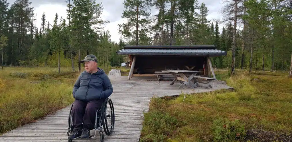 Anders tittar ut över vildmarken framför en träbod med plats för bänkar och eldstad. Han sitter i en mörkgrå rullstol, håller händerna i fickorna och är klädd i keps och varna ytterkläder. 