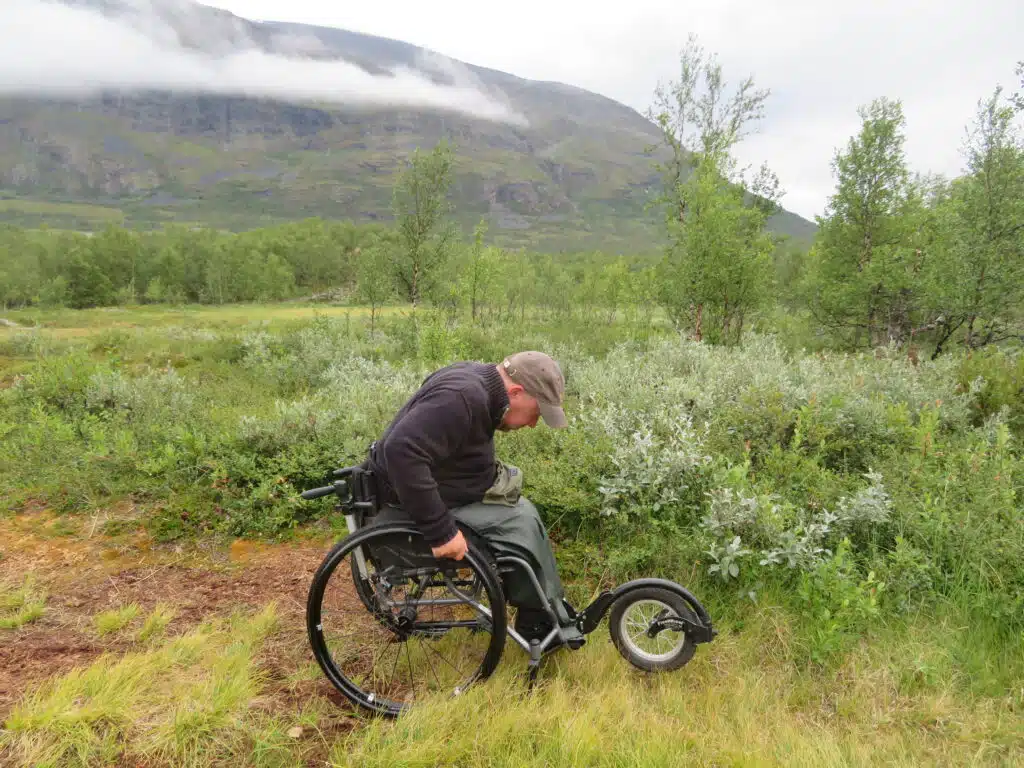 Anders pressar på rullstolens drivringar i en backe genom högt gräs. Han är klädd i en grön keps, mörklila tröja och regnbyxor