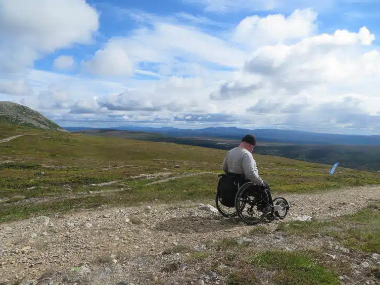 Anders tar sig fram i vildmarken med rullstol och specialanpassat framhjul. Han har en mörk mössa, ljus tröja mörka byxor och en svart rullstol. I bakgrunden syns fjälltoppar.