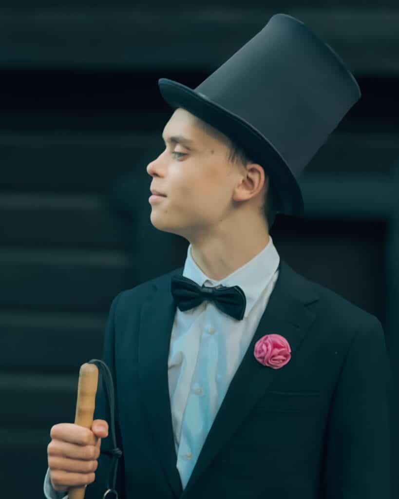 Viktor tittar åt sidan mot en mörk scenliknande bakgrund. Han är klädd i en svart hög elegant hatt, kostym, skjorta och har en rosa rosliknande utsmykning en bit ovanför bröstfickan.