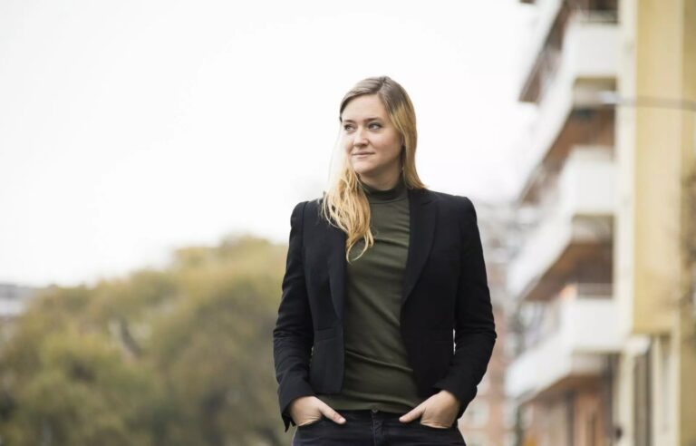 Porträttbild utomhus på Susanna. Hon står framför ett hus och grönska iklädd svart kostym med grön polotröja under. Hon tittar till vänster med ett litet leende och håller händerna avslappnat i byxfickorna.